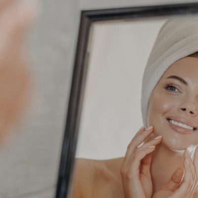 Healthy adorable young woman looks at her reflection in mirror, touches healthy glowing smooth skin